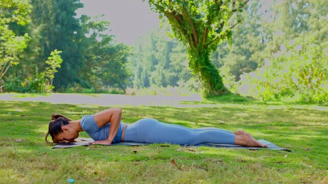 Focused young woman performs calm yoga pose on mat in lush green nature setting enjoying outdoor park doing body stretch for health fitness routine finding inner peace