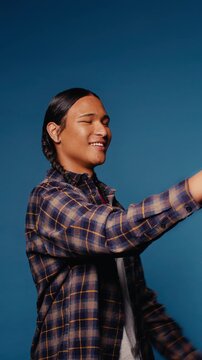 Young man smiles and uses a phone against a blue background. He takes a selfie, texts, and scrolls. Medium close-up, vertical video.