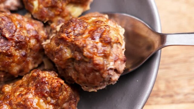 Metal spoon scoops baked meatball frikadelle from dark plate close up. Juicy roasted ground meat with browned crust