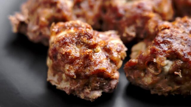 Baked meatballs with caramelized crust and melted sauce with detailed texture of cooked ground meat on ceramic black platter. Rotation