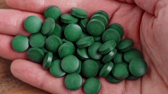 Human hand holding a heap of dark green spirulina tablets on wooden table. Pressed seaweed supplement. Dietary supplements. Top view