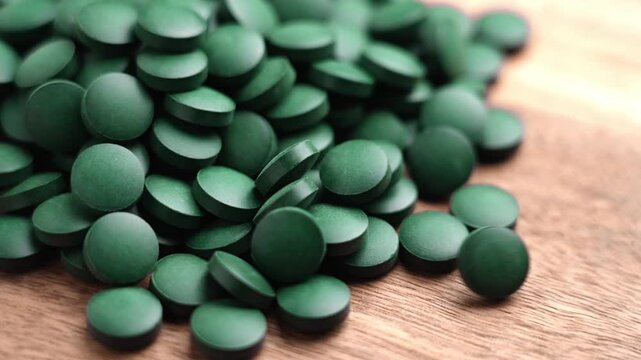 Dark green spirulina tablets falling in a heap onto a textured wooden surface. Pressed algae supplement. Detox nutrition pills. Slow motion
