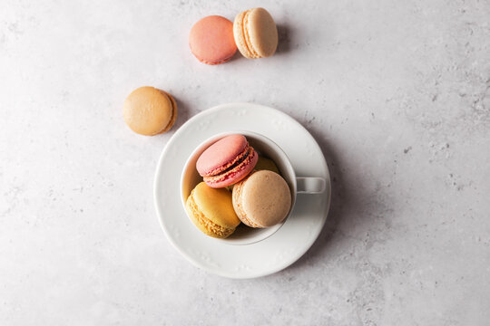 Colorful French macarons in white cup on plate with extra macarons around, horizontal top view dessert composition on gray stone background with copy space. Concept of colorful dessert background