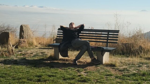 Teenage boy enjoys nature sitting on a bench high in the mountains near an Armenian church and khachkars. Person reached high mountains with silence and closeness to history and nature of Armenia.