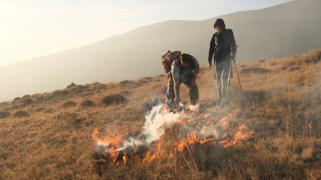 Two tourists try to extinguish a grass fire. Man pours water from plastic bottle onto flames in attempt to stop wildfire. Careless handling of fire in nature.