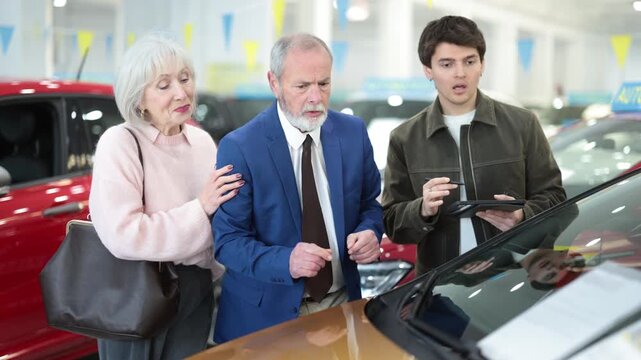  Young car salesman presents car to mature man and woman at car dealership, discussing price and terms of sale