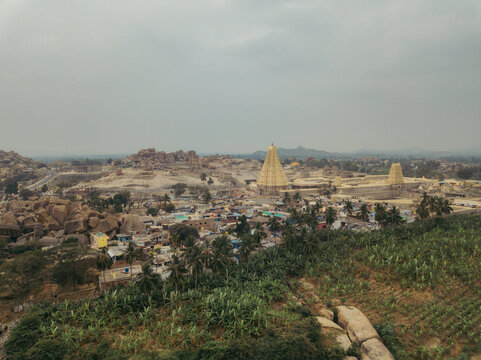 Aerial view of the ancient Virupaksha Temple's towering gopurams piercing the skyline amidst the rocky terrain and verdant fields, Hampi, Karnataka, India.