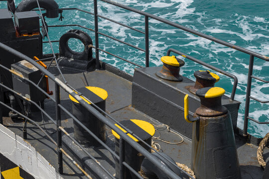 Mooring bollards on a ferry deck with yellow markings and metal railings, set against turquoise sea waves, highlighting maritime equipment and nautical transport environment.