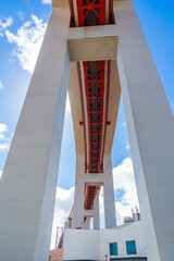 Obraz premium Low angle view from beneath a large modern bridge showing massive concrete pillars and red steel framework against blue sky with clouds.