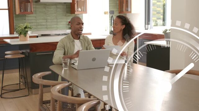 Couple at kitchen island sorting bills, pointing at laptop and papers as animated clock expands