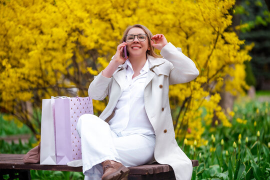 Woman talking on smartphone on bench with shopping bags. Spring leisure and lifestyle.