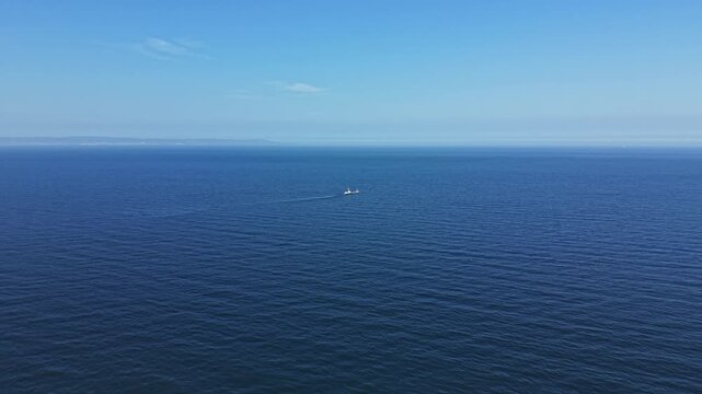 A boat moves slowly over the surface of an expansive ocean. The water appears calm and blue with a clear sky above during daytime. There is no land in sight.