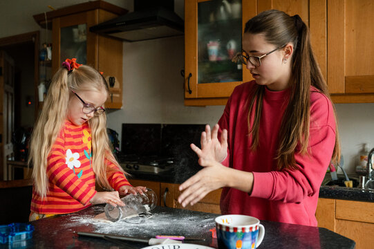 Child rolling out pastry with a glass while her teenage sister rubs off flour from her hands