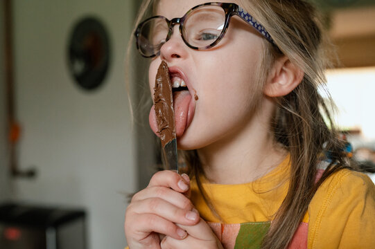 Child with glasses licking chocolate spread off a knife