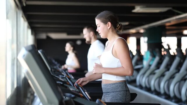 Girl in gym runs with help of treadmill. She does exercises on simulator and monitors body indicators on screen. Girl doing exercise in health club, perform sport routine