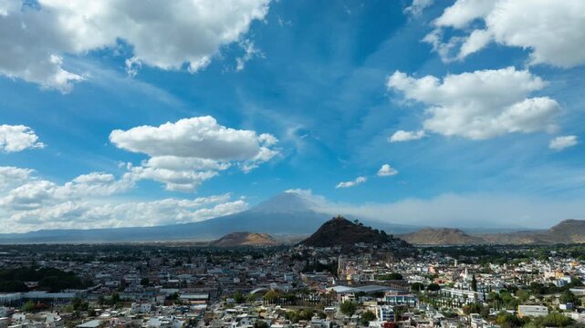 Aerial hyperlapse push in over downtown Atlixco, Puebla, Mexico, showing a closer urban view with Cerro de San Miguel, city landmarks and Popocatepetl volcano under a blue sky with fast moving clouds.