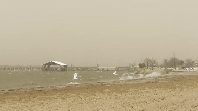 Rough waves crash onto a sandy beach while blowing dust fills the air. A distant pier stands against the turbulent lake.