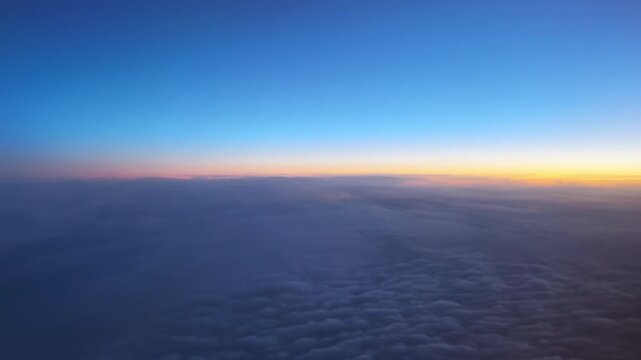 Aerial shot above a cloud layer showing blue sky and a warm orange sunrise glow across the horizon.