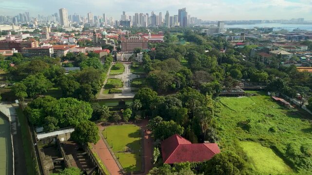 Aerial drone shot reveals lush greenery of Fort Santiago park within historic walled city of Intramuros Manila, showing red rooftops and Pasig River against modern metropolitan horizon