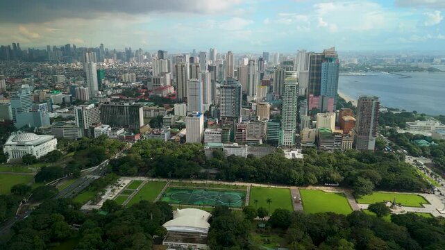 High aerial shot over Rizal Park lush green lawns and fountain lagoon revealing sprawling Manila cityscape, skyscrapers and Manila Bay coastline