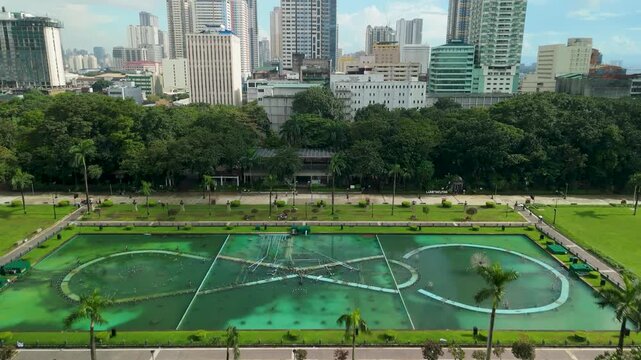 Drone tilts up from iconic Rizal Park fountain lagoon and auditorium canopy roof revealing Manila urban skyline contrasting lush green park trees
