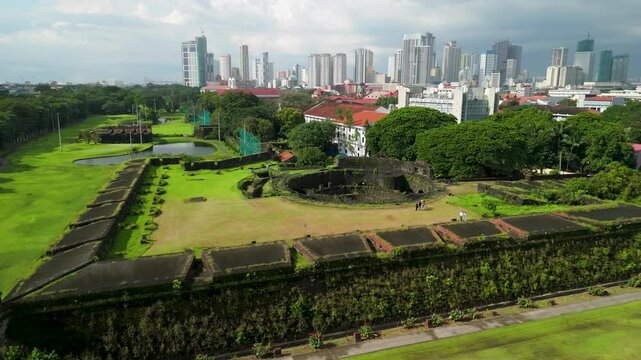 Drone parallax circles Baluarte de San Diego revealing moss-covered stone bastion courtyard with tourists, colonial buildings and Manila skyline beyond