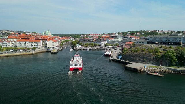 Drone follows red and white catamaran passenger ferry carrying tourists as it enters Stromstad harbor channel with colorful city and rocky coastline in background
