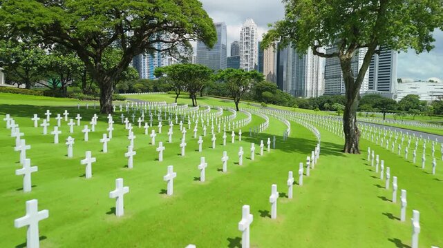 Aerial flyover above white marble crosses in circular rows at Manila American Cemetery with Bonifacio Global City skyscrapers in background under bright sunny sky