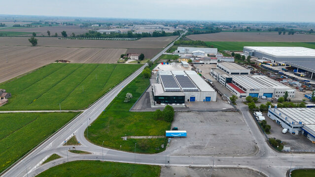 Industrial zone along Via Emilia SS9 near the A1 exit of Fiorenzuola d Arda in the Po Valley with warehouses, service roads and truck activity integrated within surrounding agricultural land