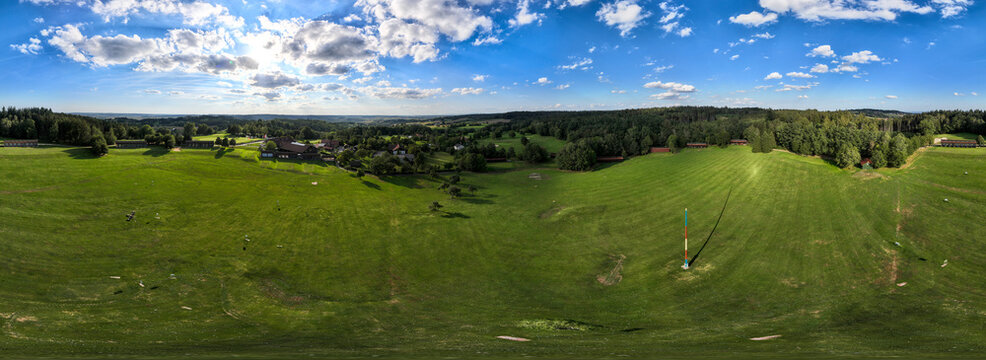 Aerial view of a vibrant green landscape dotted with trees and buildings, under a bright blue sky with fluffy clouds, Bad Griesbach, Germany.