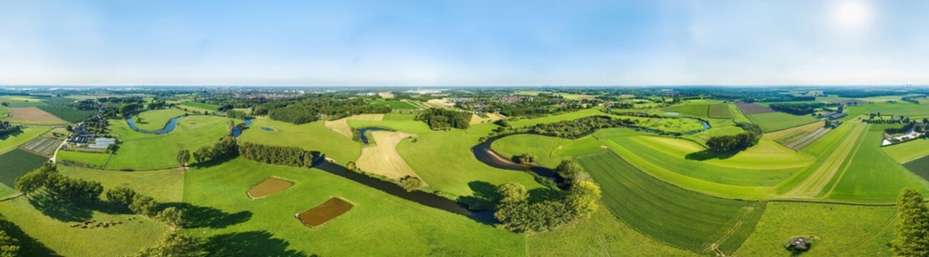 Aerial view of meandering river Roer cutting through vibrant green meadows and cropland in front of villages, in 360 degrees panorama, valley Roerdal, Limburg, Netherlands.