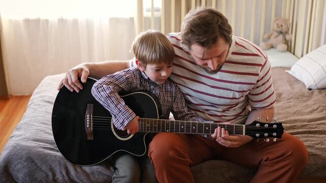 Handsome dad teaches his cute son to play the guitar sitting on sofa in the living room at home. Happy Father's Day. Father teaching his child to play guitar. Kid Learning music skill. Art education