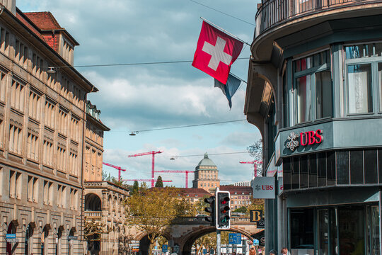 Zurich, Switzerland, April 16, 2026. A street-level view of a UBS bank branch with a Swiss flag, leading toward the University of Zurich dome and construction cranes in the distance under a bright sky
