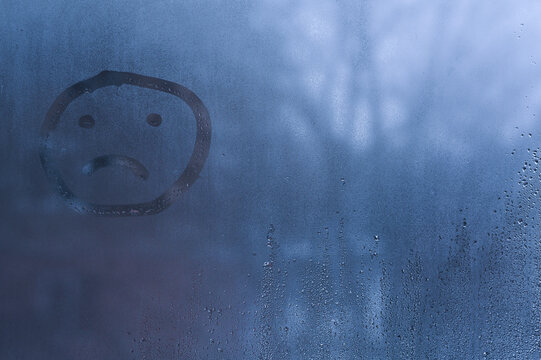 Sad face symbol drawn on condensation covered glass, finger line forming simple unhappy expression. rueful face appearing on foggy window surface with water drops, emotion concept, loneliness feeling,