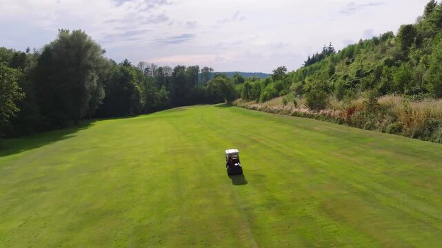 Aerial view of a golf cart on a lush green golf course, contrasting with the darker green trees and cloudy sky, Bad Griesbach, Germany.
