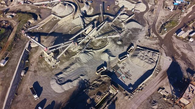 Heavy machinery moves raw materials around a construction site filled with sand and gravel. Various structures are visible, showcasing an active industrial scene amidst natural surroundings.