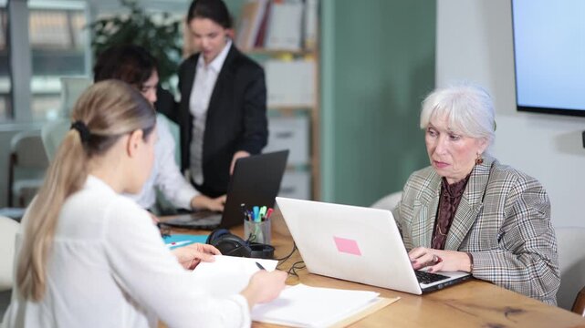 Mature woman sitting at desk is working on laptop computer among staff colleagues. Working day in office of business structure, manager is working at computer. 