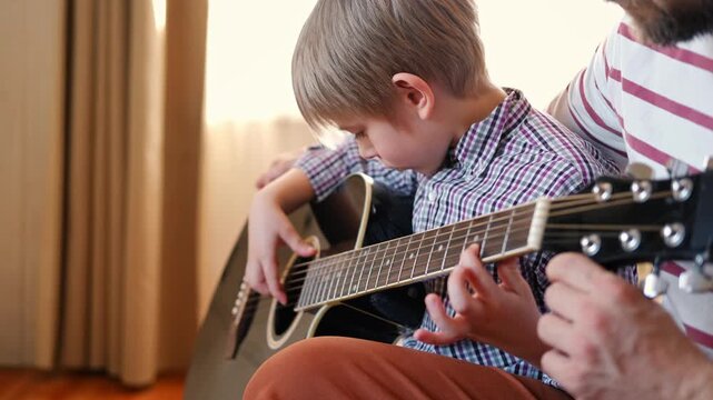 Handsome dad teaches his cute son to play the guitar sitting on sofa in the living room at home. Happy Father's Day. Father teaching his child to play guitar. Kid Learning music skill. Art education