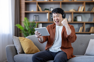Obraz premium Asian man with headphones celebrating success and winning a video game on a digital tablet, sitting on a couch in a cozy home living room, expressing excitement and achievement