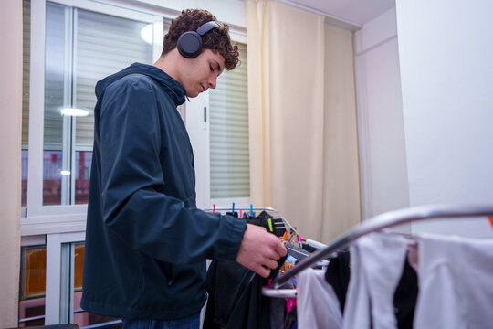 Young man doing laundry, hanging clothes on a drying rack while listening to music with headphones at home