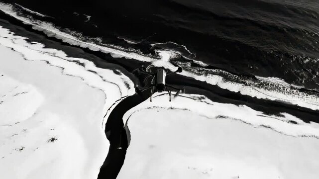 Hovering quadcopter drone rotating and advancing over snowy coast, monitoring dark stream and surf