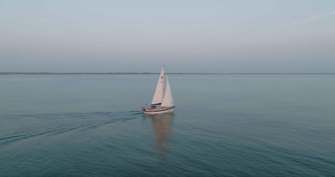 Aerial view of a lone sailboat with white sails cutting through the calm, blue waters under a pale sky, Egmond aan Zee, Netherlands.