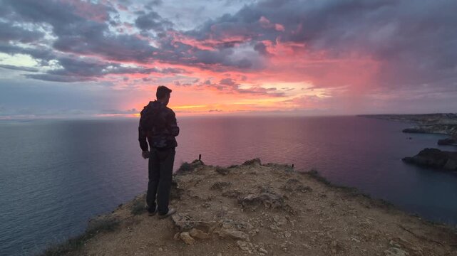 Man, cliff, sunset, dramatic sky reflecting pink and purple light over the vast ocean view