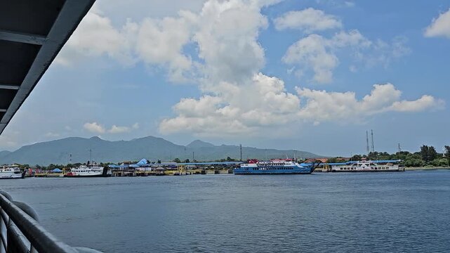 Video from a sea vessel approaching Gilimanuk Harbor Bali, showing nearby ships, calm water surface, distant mountains, port structures, and clear sunny sky.