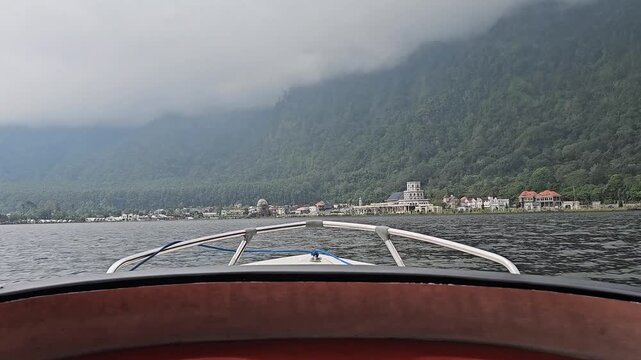 Front POV from a speedboat on Lake Beratan showing boat bow, misty mountains ahead, distant houses along the shoreline, calm water surface, and light morning fog.