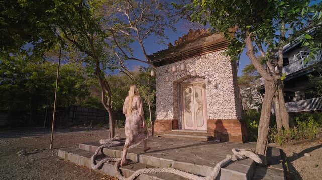A woman walks toward a gate made of coral stone walls in a tropical area. The scene highlights cultural architecture, warm sunlight, and a peaceful travel atmosphere with natural textures.