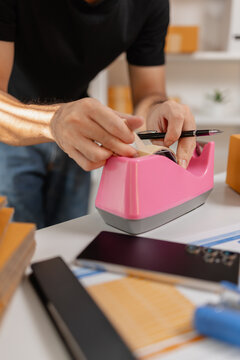 Small business owner sealing parcel with tape dispenser while preparing packages for shipment, organizing delivery orders and packaging process, representing ecommerce and logistics workflow.