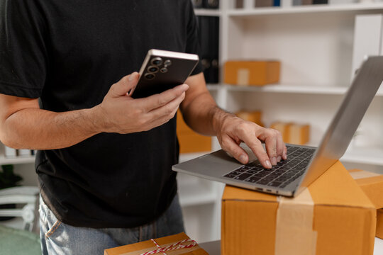 Small business owner typing on laptop while checking orders on smartphone, managing ecommerce sales and shipping tasks, representing digital business workflow and online order processing.