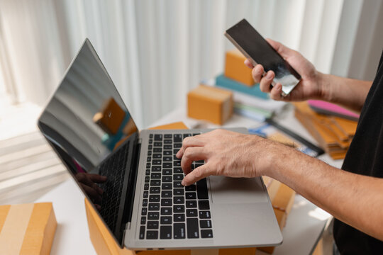 Small business owner typing on laptop while checking orders on smartphone, managing ecommerce sales and shipping tasks, representing digital business workflow and online order processing.