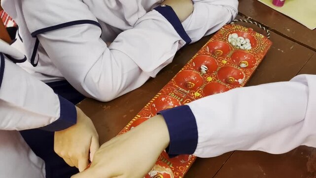 Uniformed Students Playing Traditional Congkak Mancala Board Game At Wooden Table